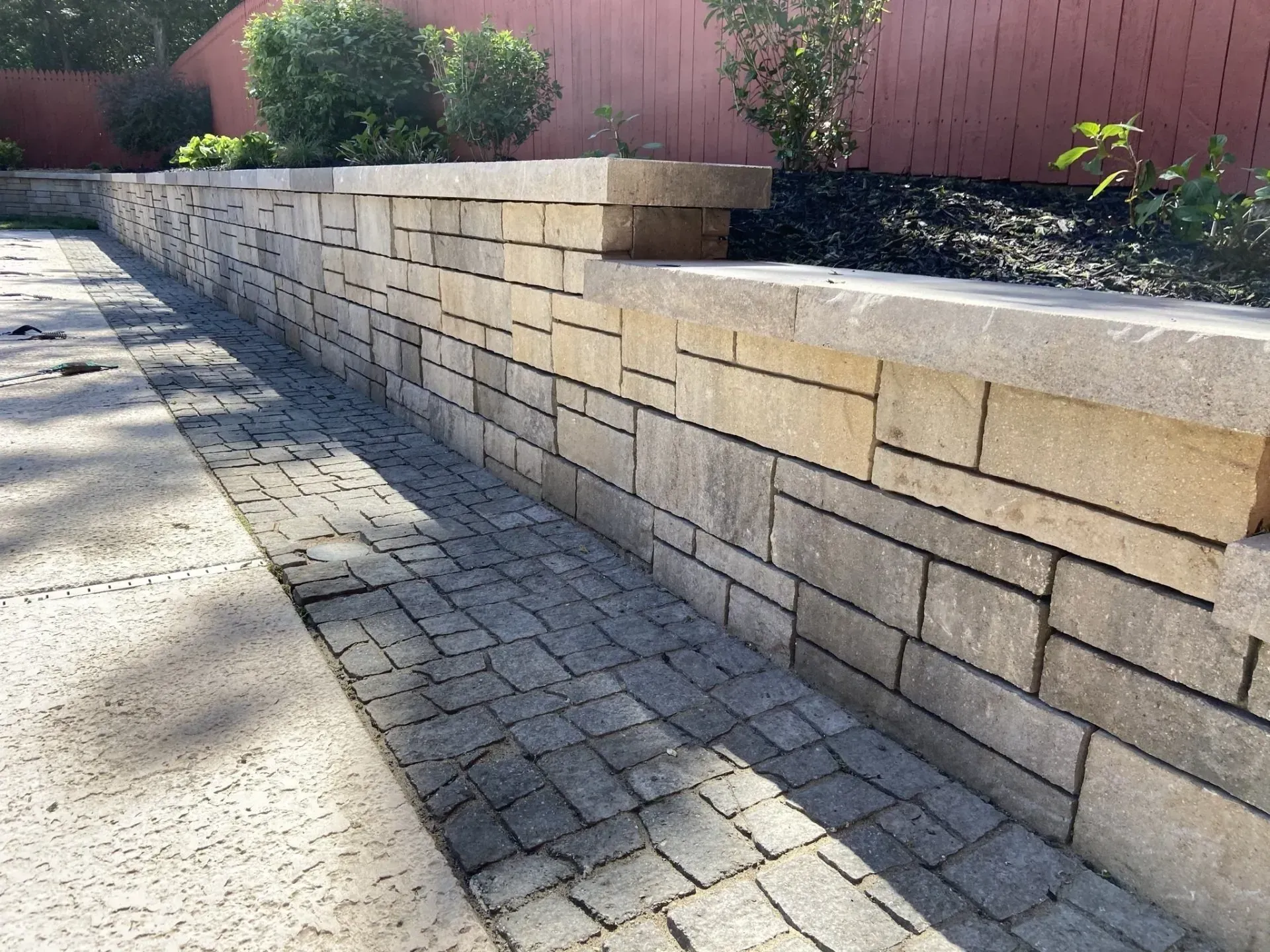A stone retaining wall alongside a patio made of gray square pavers, with dark mulch and plants behind the wall.