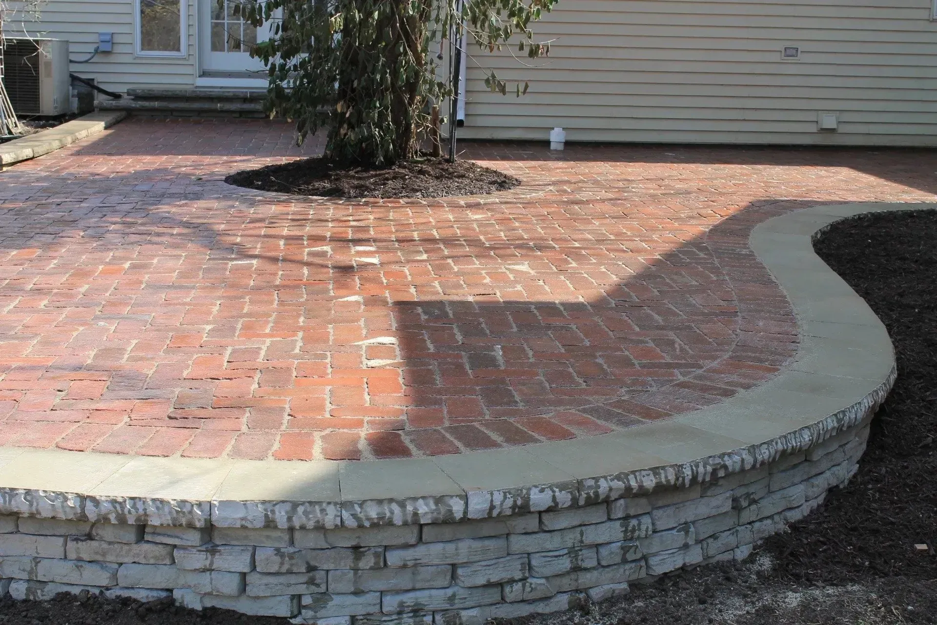 A curved brick patio with a low, light-colored stone retaining wall in a backyard setting.
