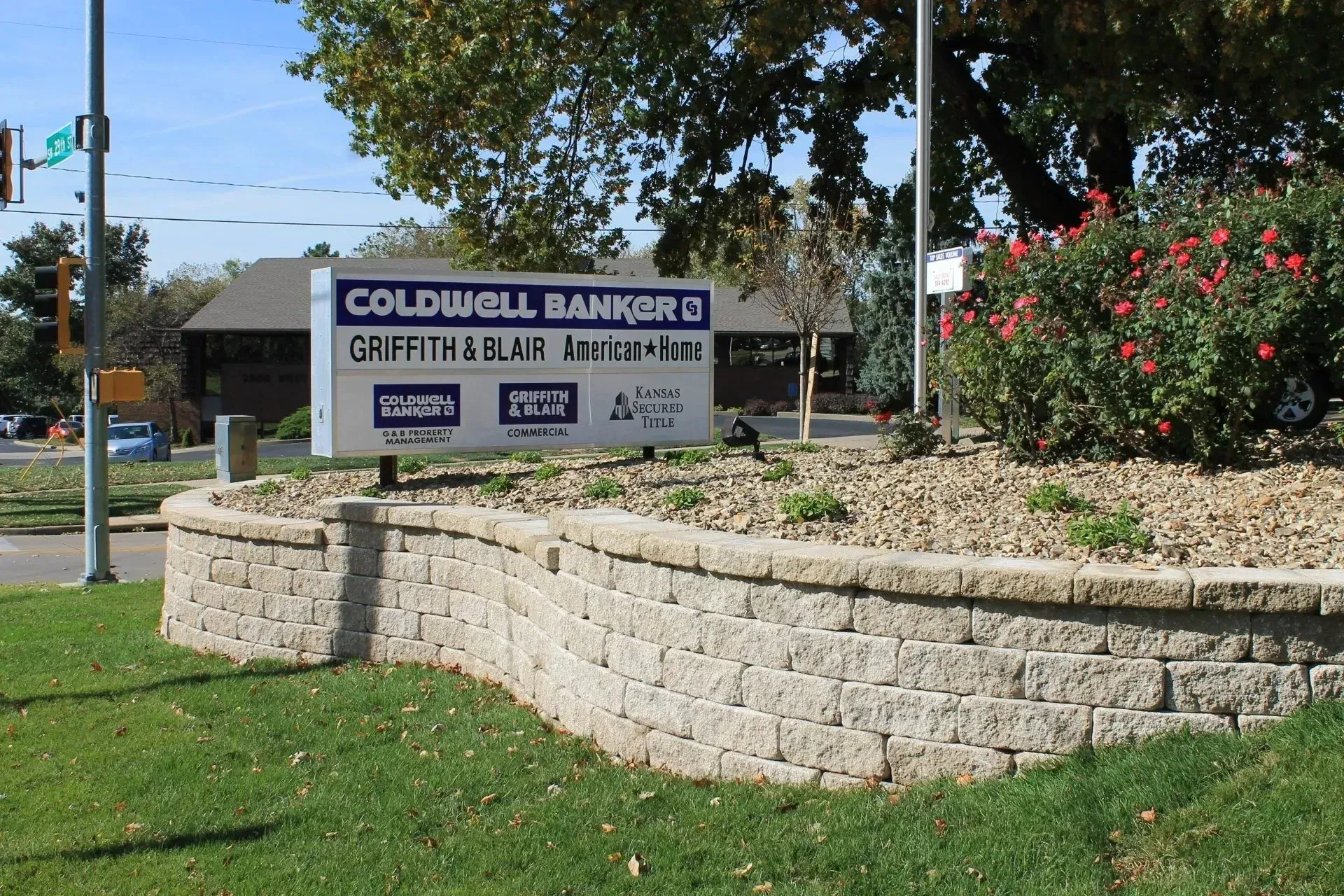 A Coldwell Banker real estate office sign stands behind a tan stone retaining wall on a grassy lawn with trees and shrubs.