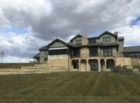 A two-story grey-sided house with a stone base, arched lower-level openings, and a large lawn under a cloudy sky.