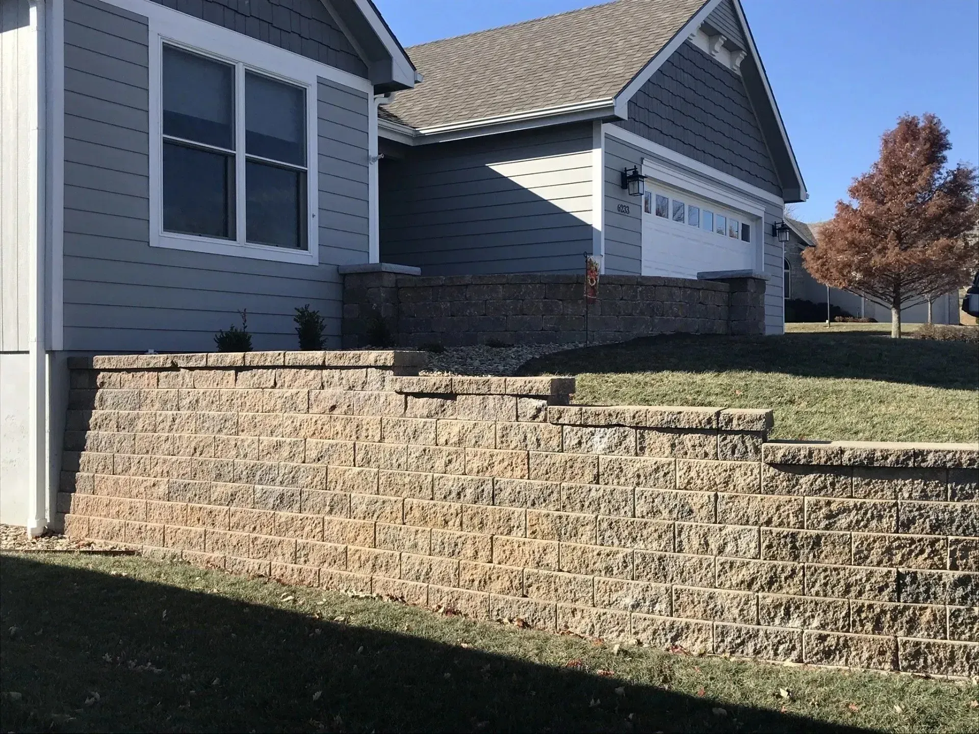 A gray suburban house with a large, tiered brown stone retaining wall in the front yard under a clear blue sky.