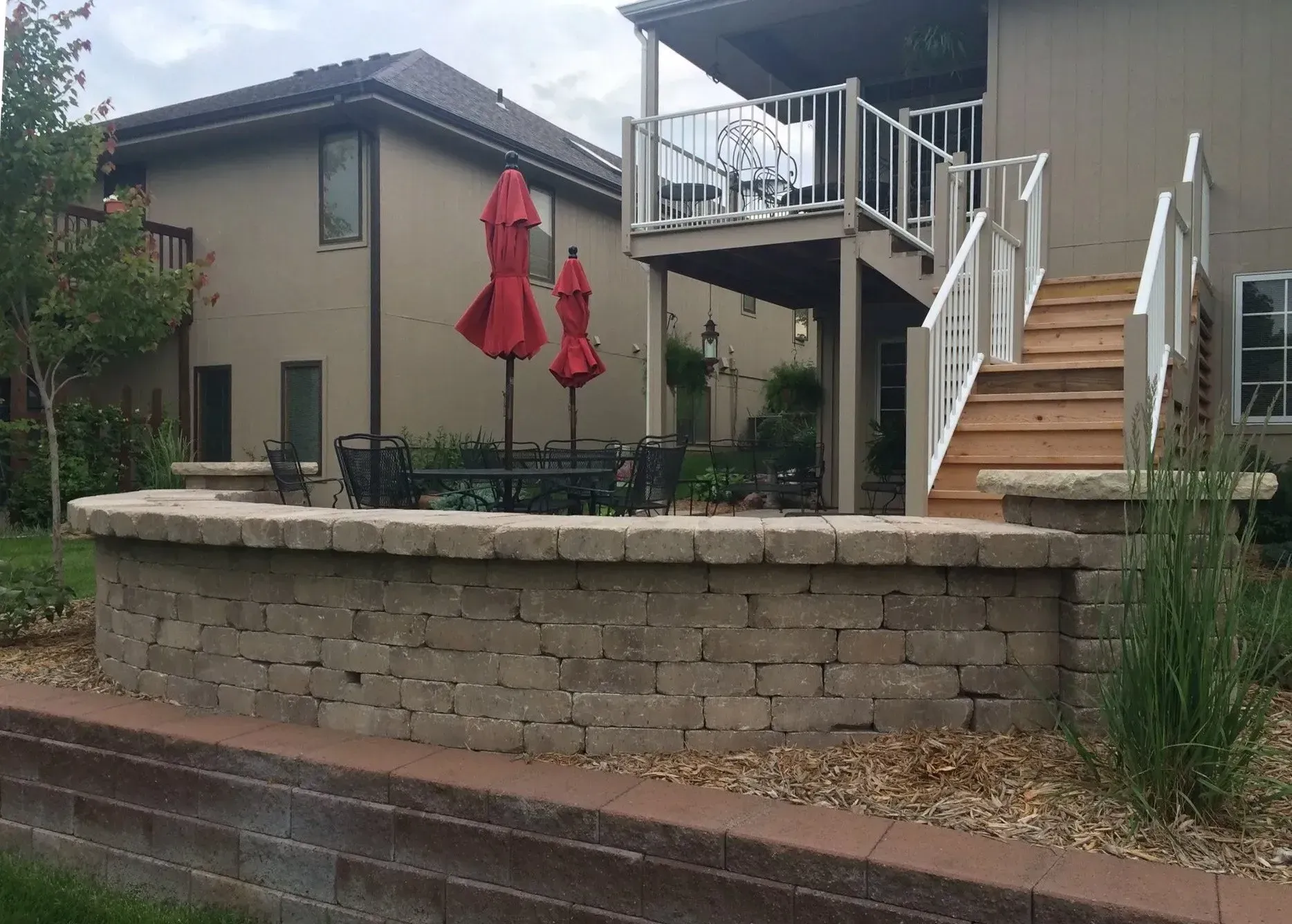 A low, curved stone retaining wall in front of a house patio with red umbrellas, wooden stairs, and a raised deck.