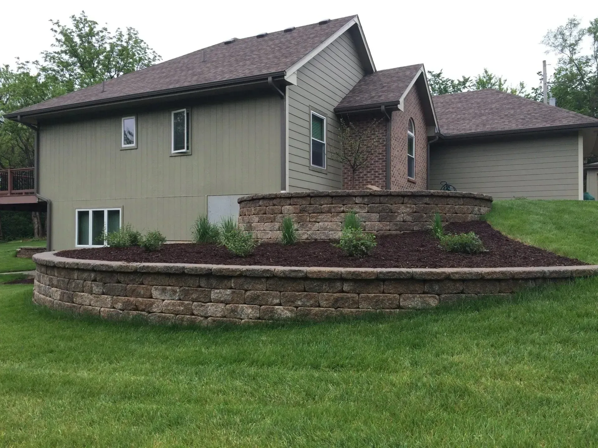 A tan two-story house with a brick accent features a two-tiered, curved stone retaining wall in the grassy front yard.