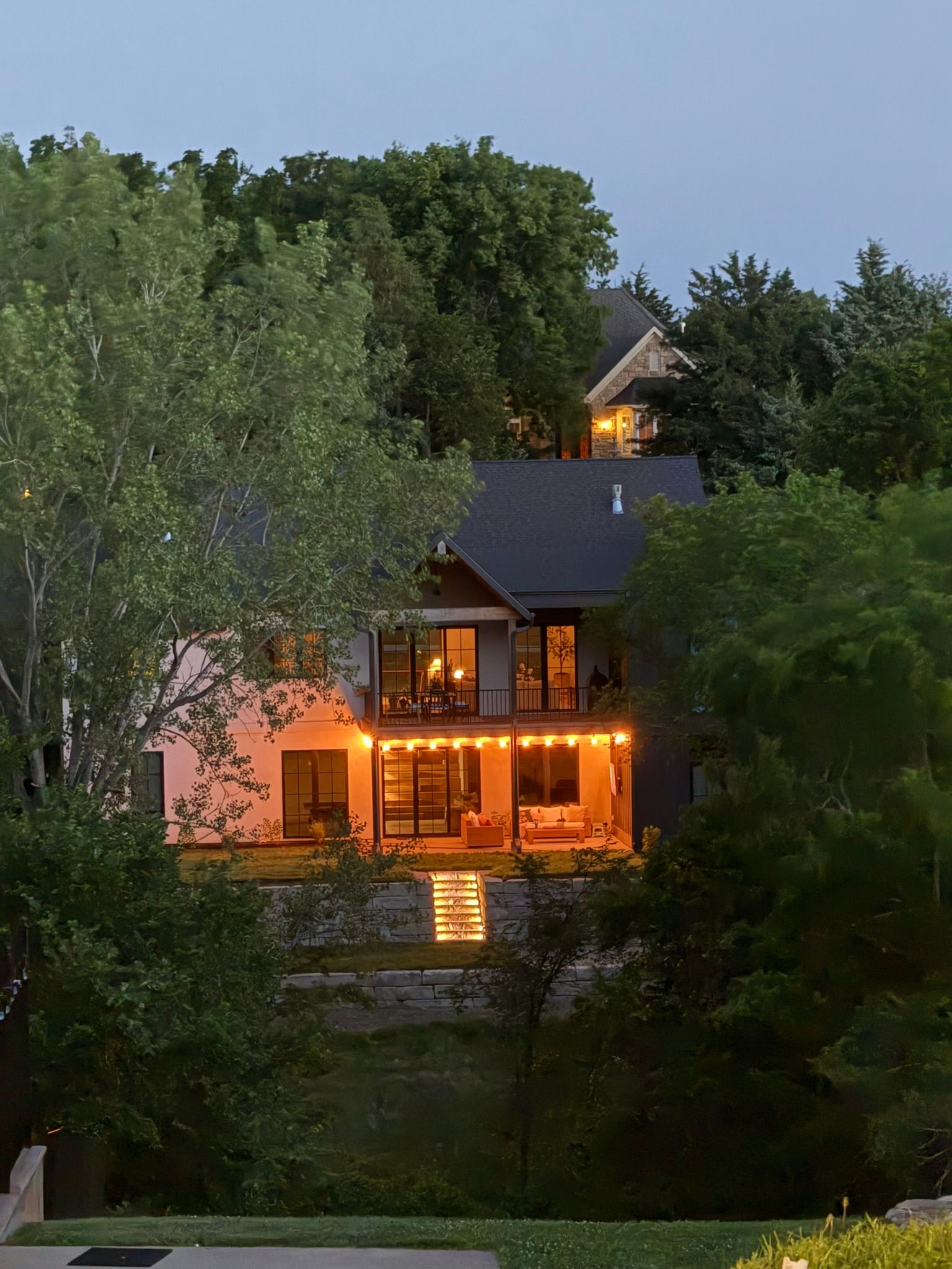 A multi-story house at dusk, illuminated by warm string lights and interior lamps, nestled amidst lush green trees.