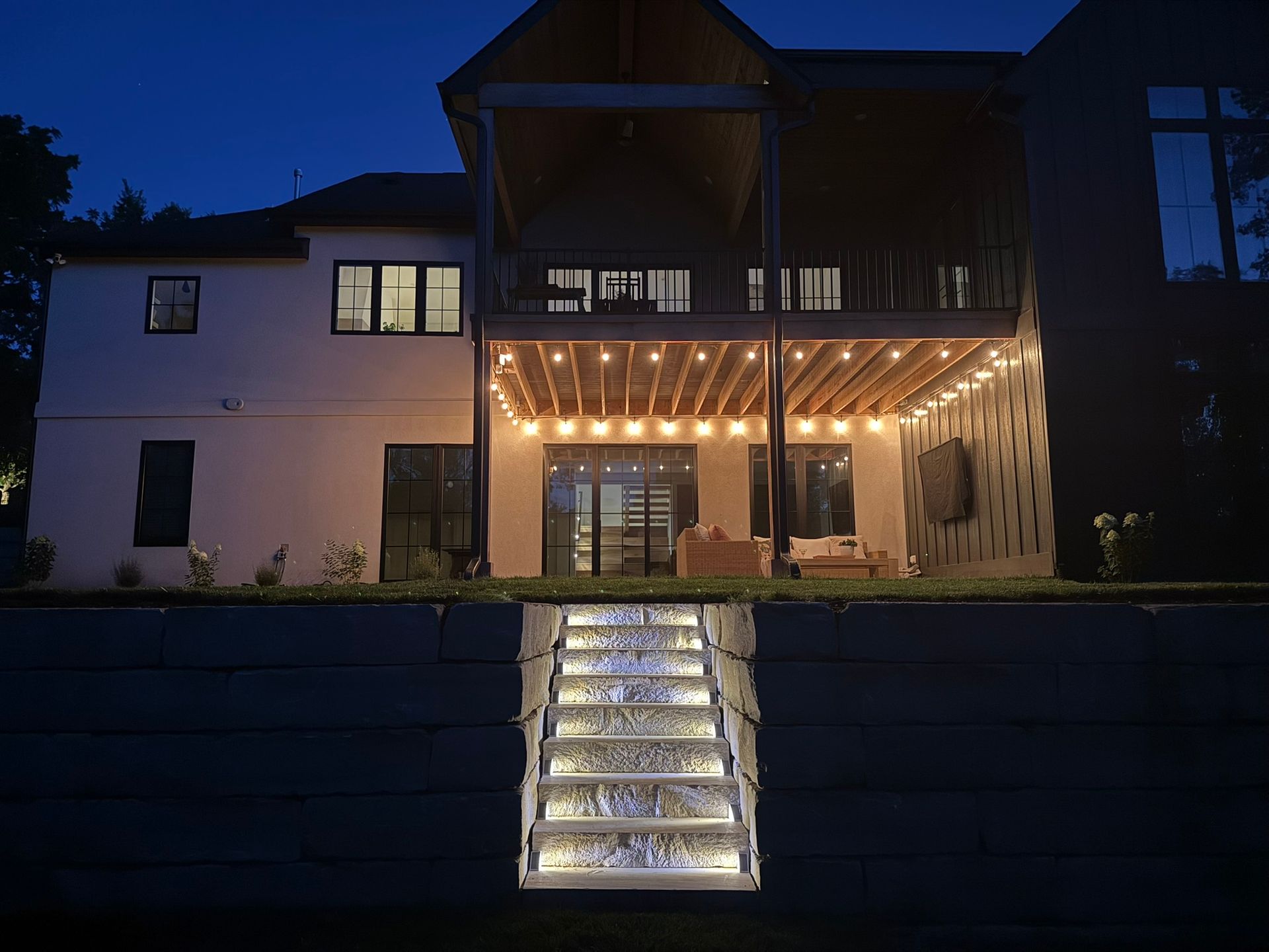 A brightly lit stone staircase set into a dark retaining wall, leading up to a two-story house with illuminated deck lights.