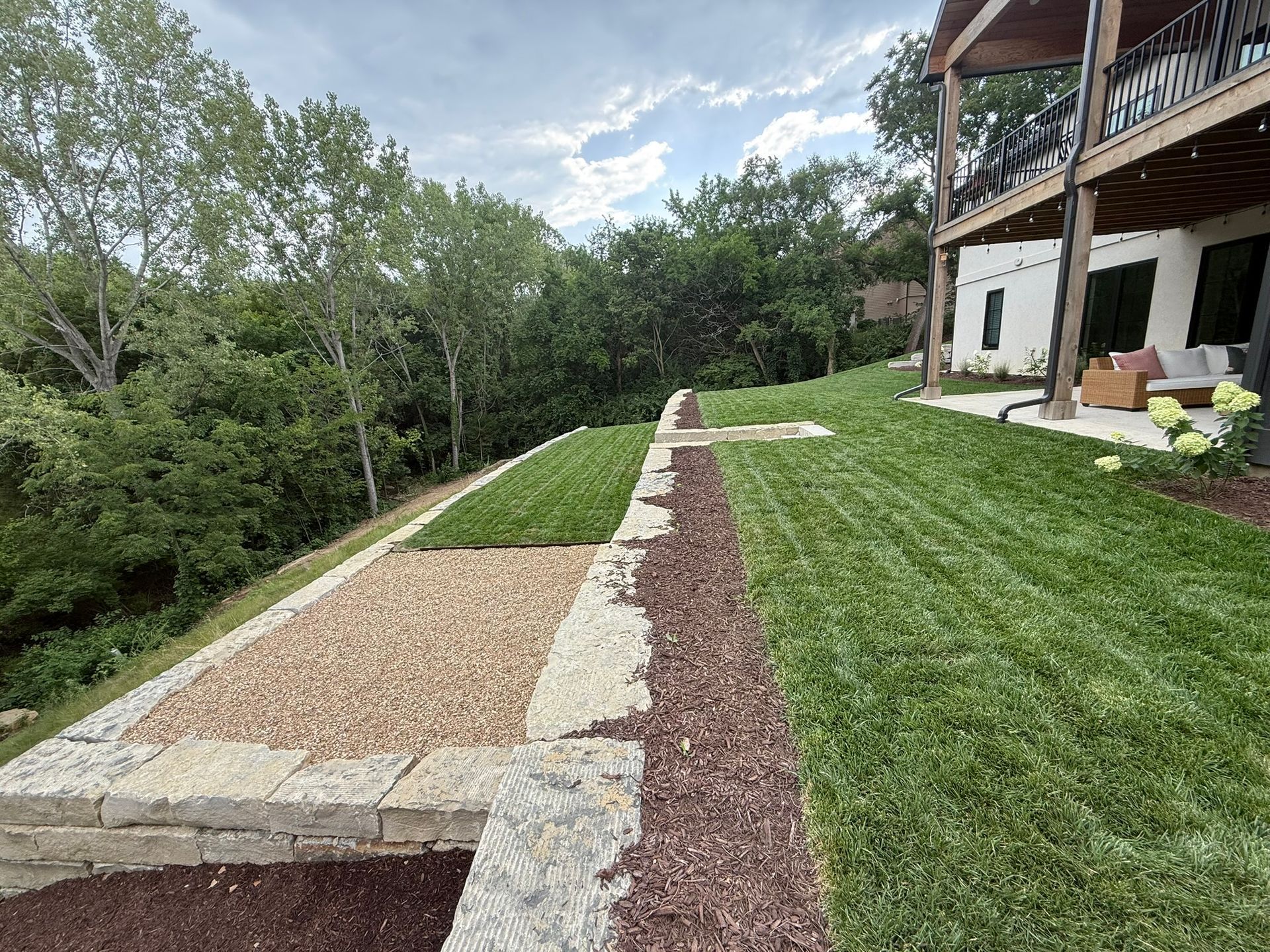 A terraced backyard landscape with stone retaining walls, patches of grass, gravel, and mulch, next to a house patio.