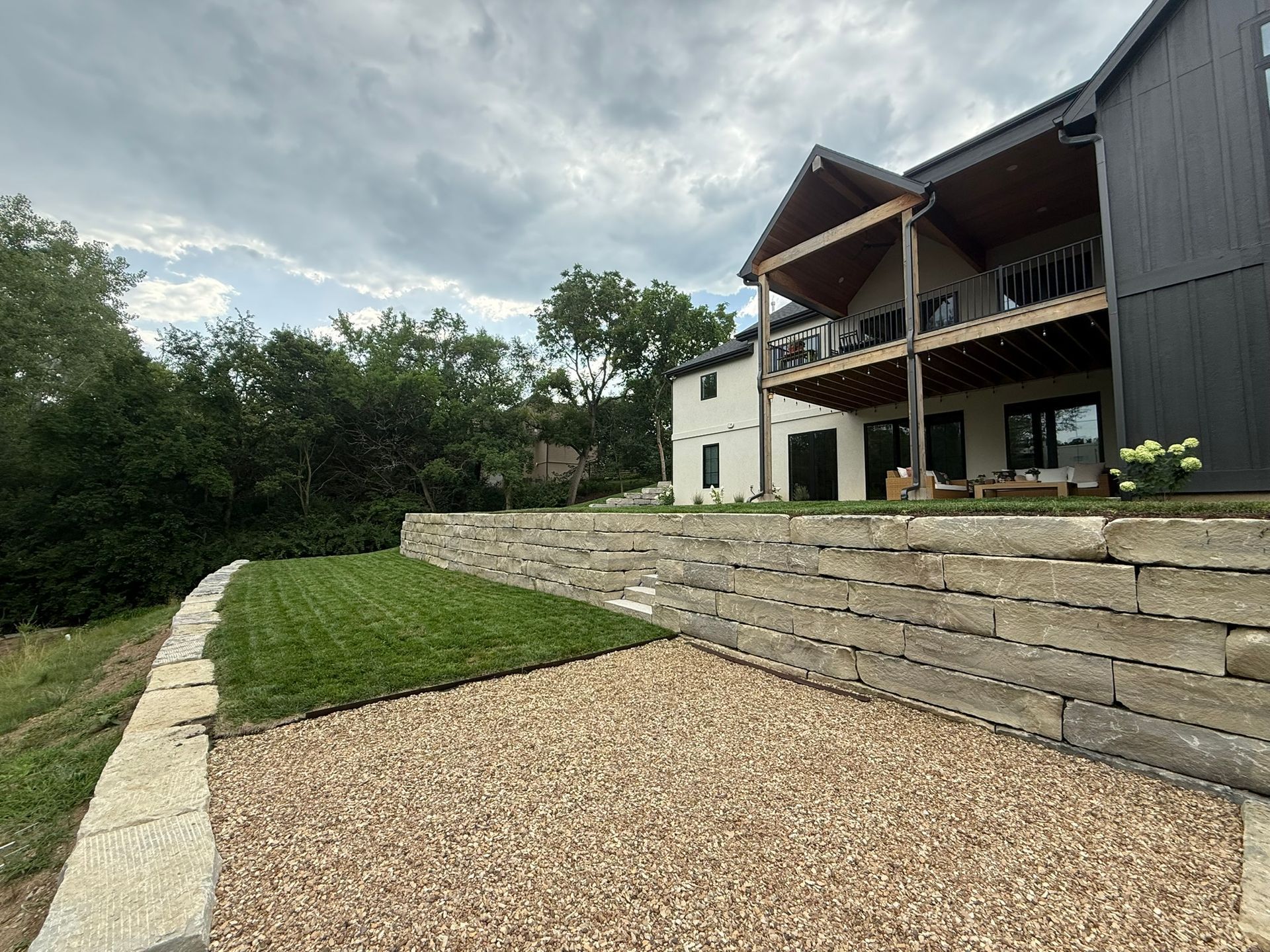 A landscape view of a modern house with a large stone retaining wall separating a gravel patio from a raised grassy yard.