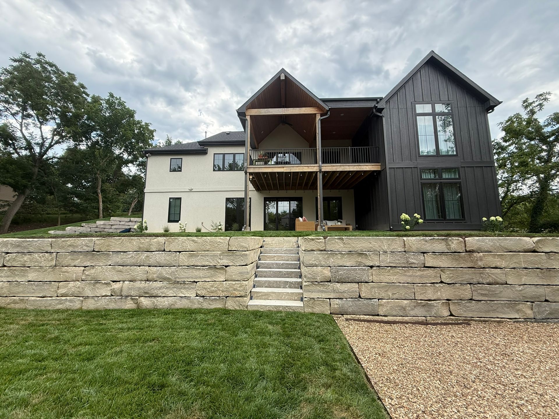 Two-story house with white stucco and black siding, featuring a central covered balcony and tiered stone retaining wall.
