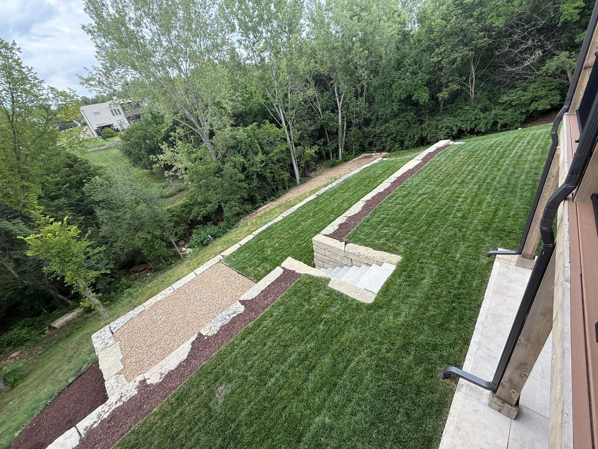 Tiered backyard landscape with stone retaining walls, a gravel patio, lawn, and surrounding trees.