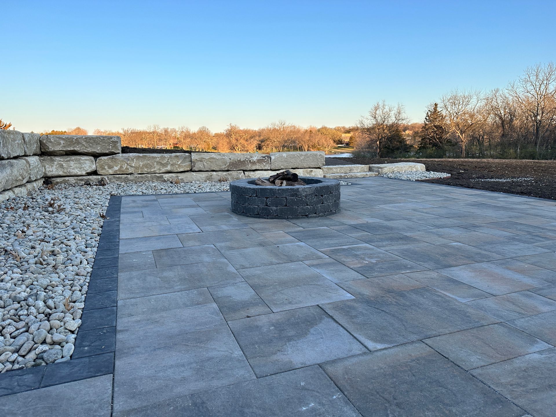 A stone patio with a central circular fire pit, bordered by white gravel and a retaining wall under a clear blue sky.