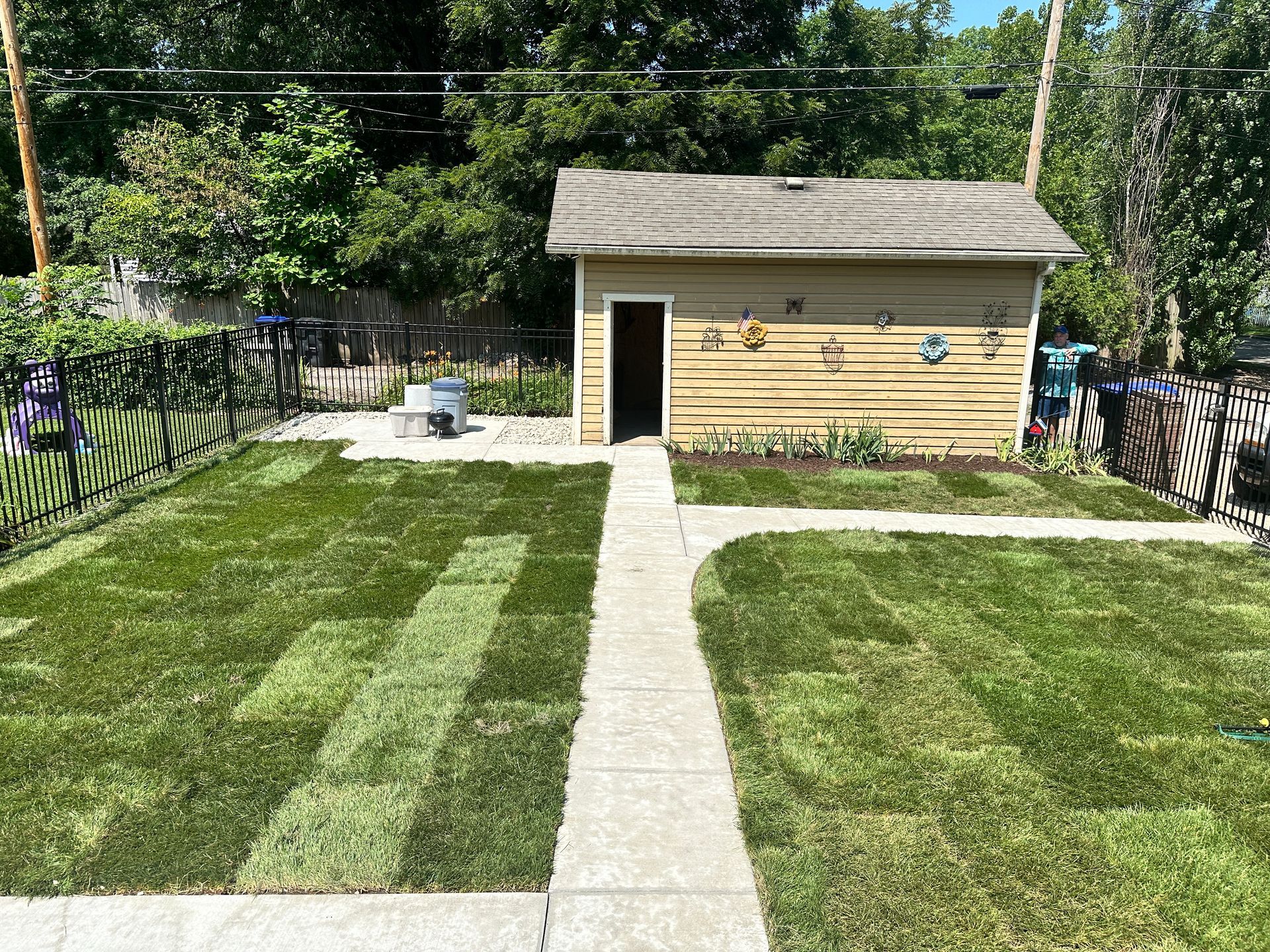 A concrete walkway leads through a freshly sodded backyard toward a tan garage with metal wall decorations.