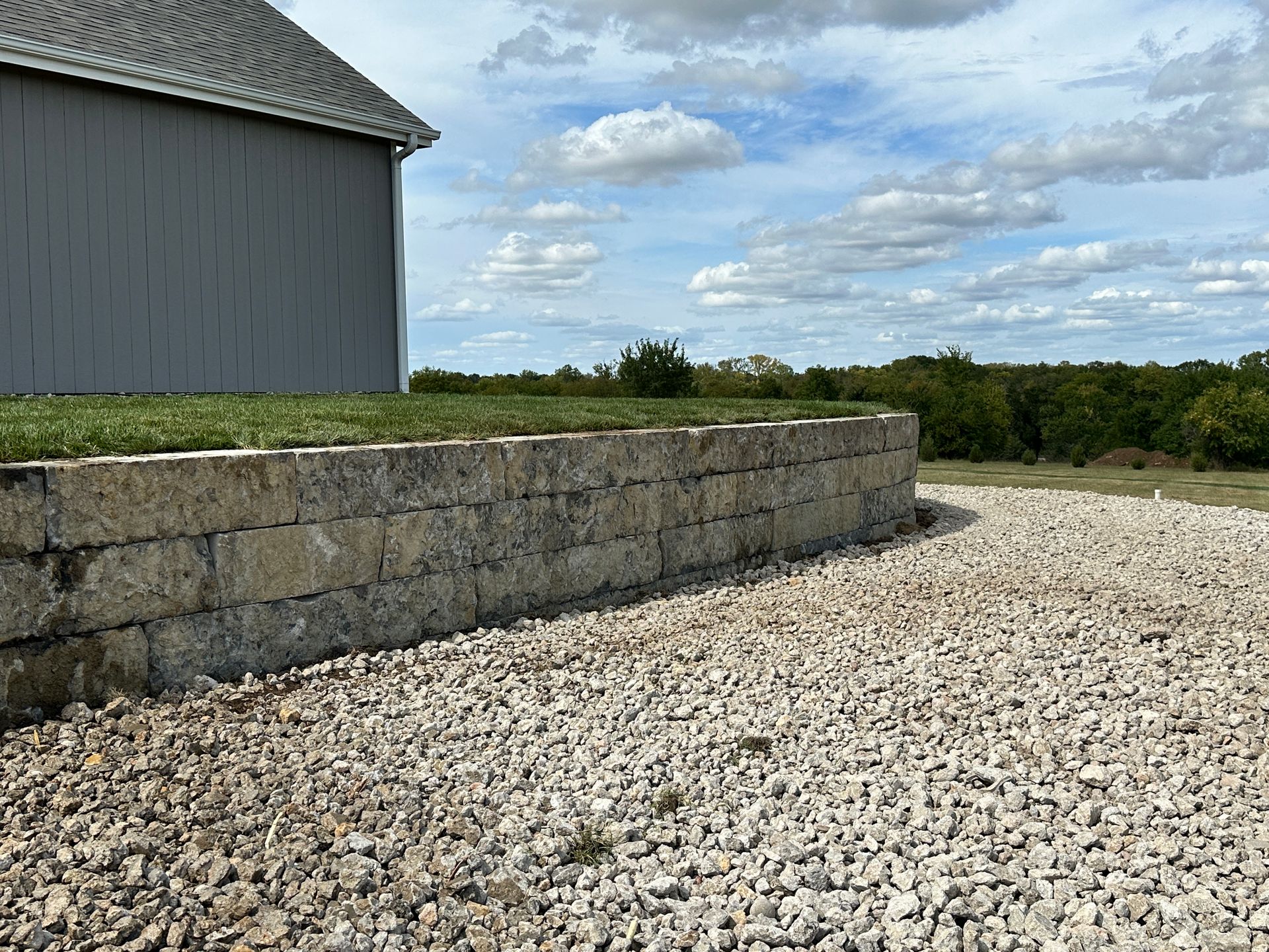 A stone retaining wall separates a grassy yard from a graveled area next to a building under a partly cloudy sky.
