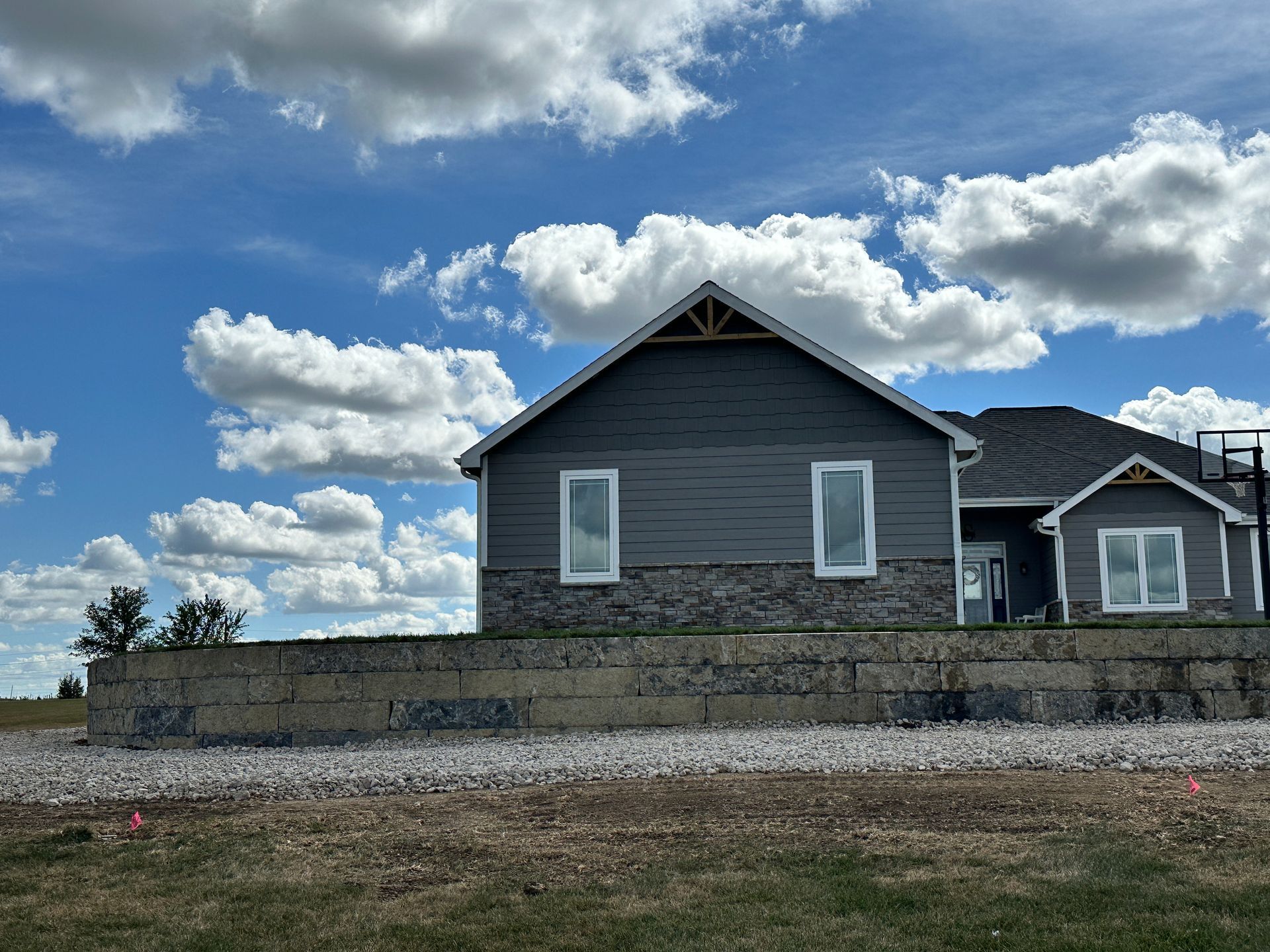 A single-story house with dark grey siding and stone accents sits behind a long, light-colored concrete retaining wall.