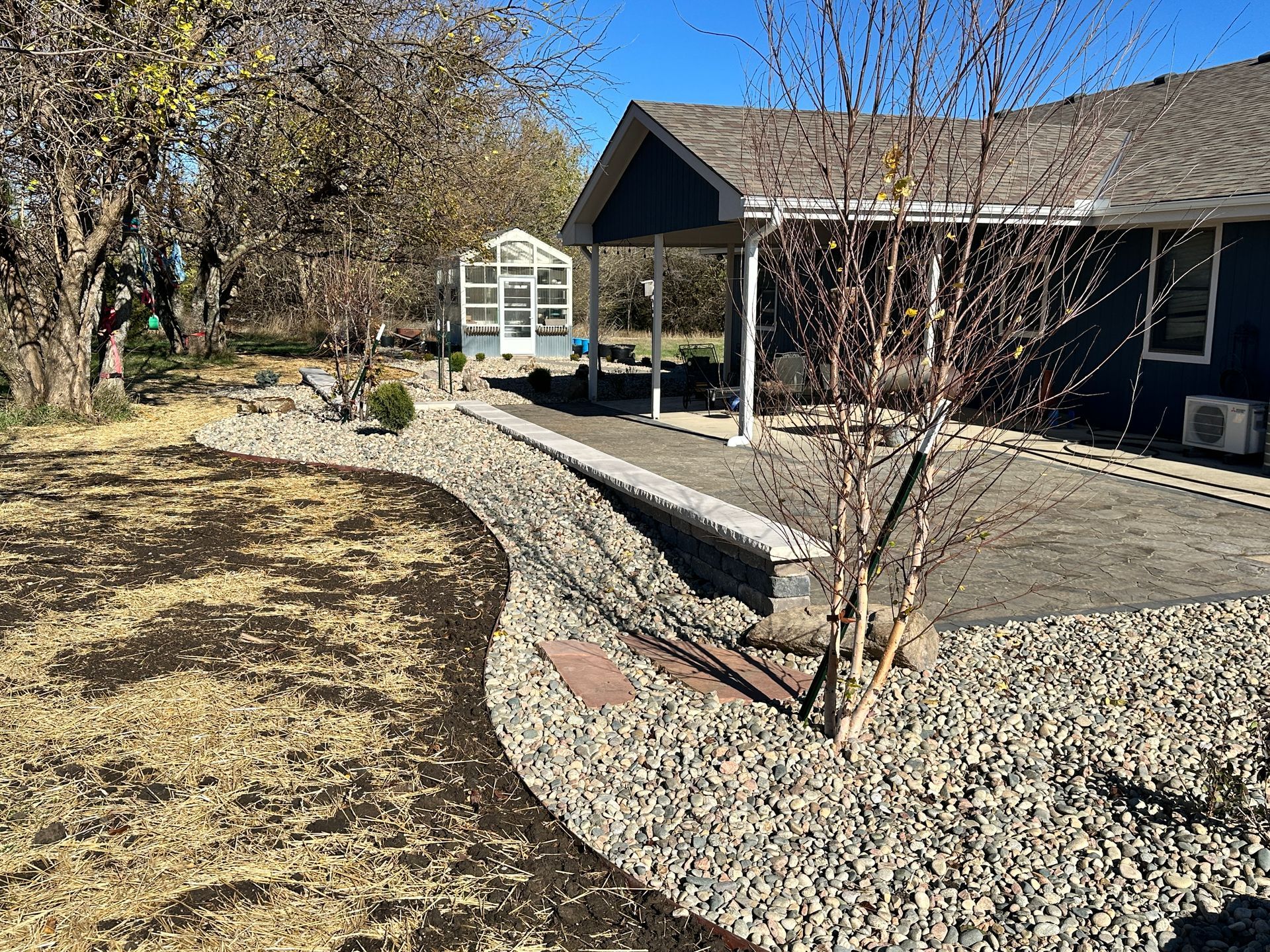 A view of a backyard patio with a stone retaining wall, gravel landscaping, a small greenhouse, and trees under a blue sky.