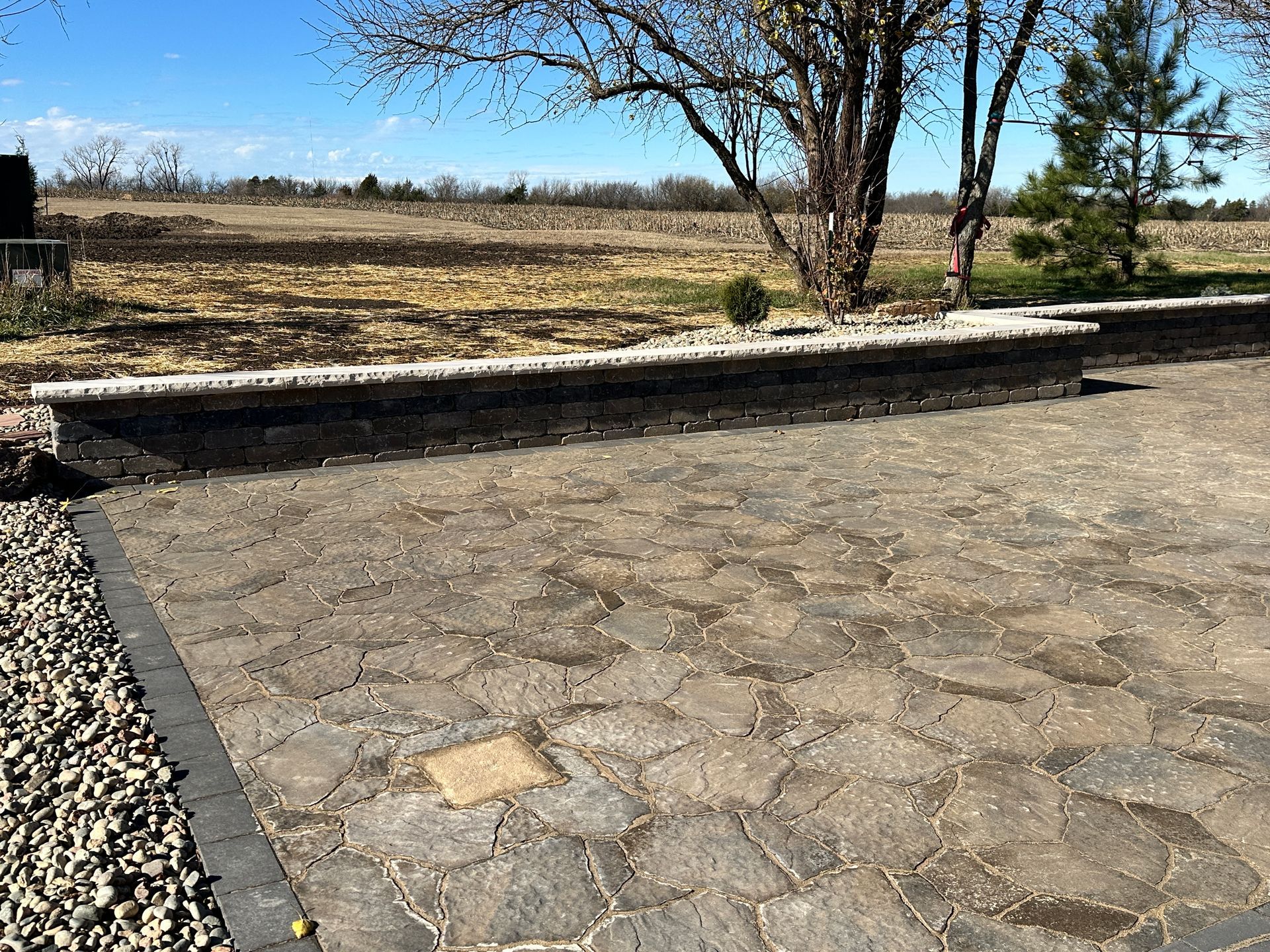 A stone patio with a low retaining wall, white gravel edging, and a field in the background under a clear blue sky.