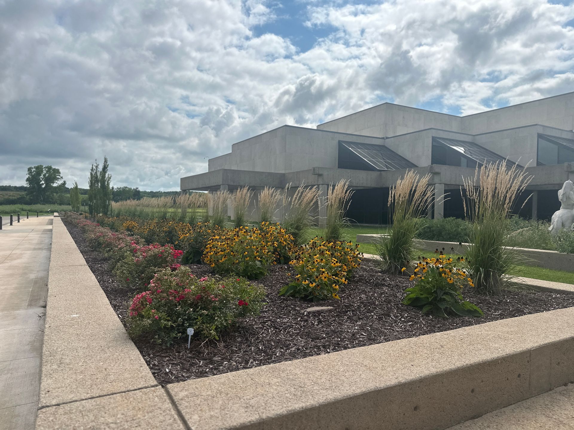 Modern concrete building with a large planter featuring colorful flowers and ornamental grasses under a cloudy sky.
