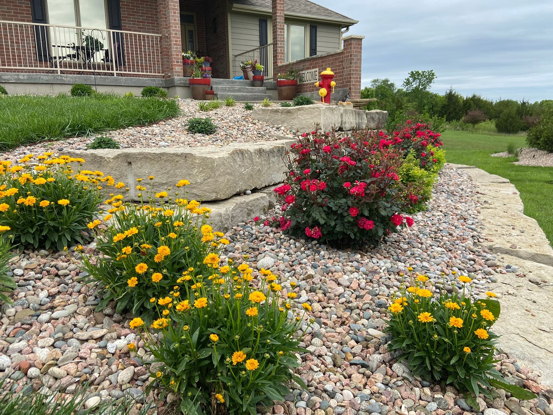 A landscaped front yard featuring tiered limestone retaining walls, gravel mulch, yellow perennials, and a rose bush.