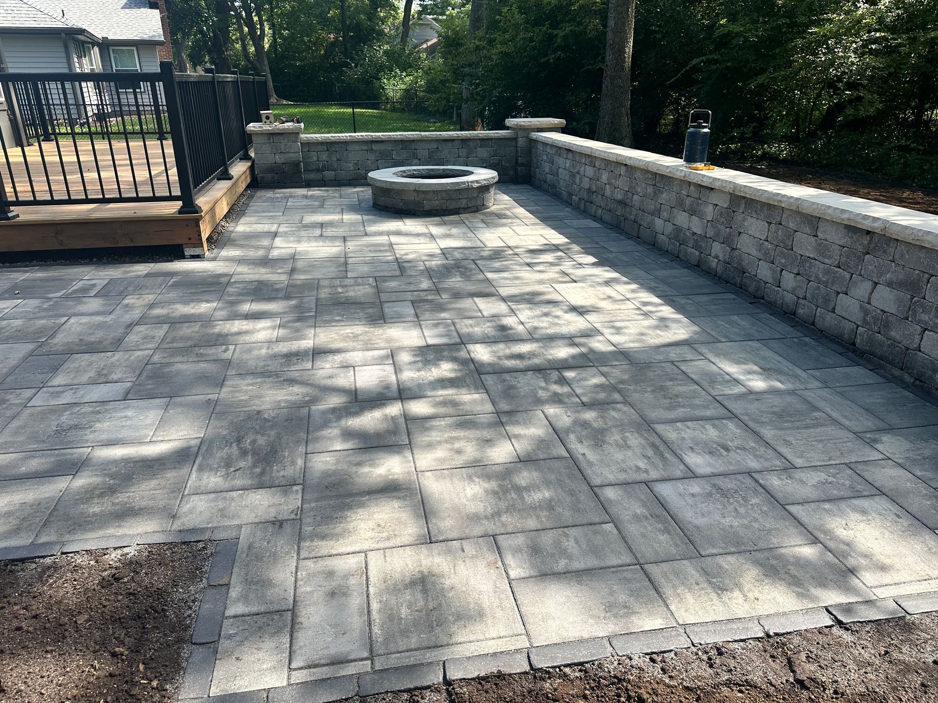 A backyard patio featuring a stone fire pit, a raised stone retaining wall, and a raised wooden deck with a black railing.