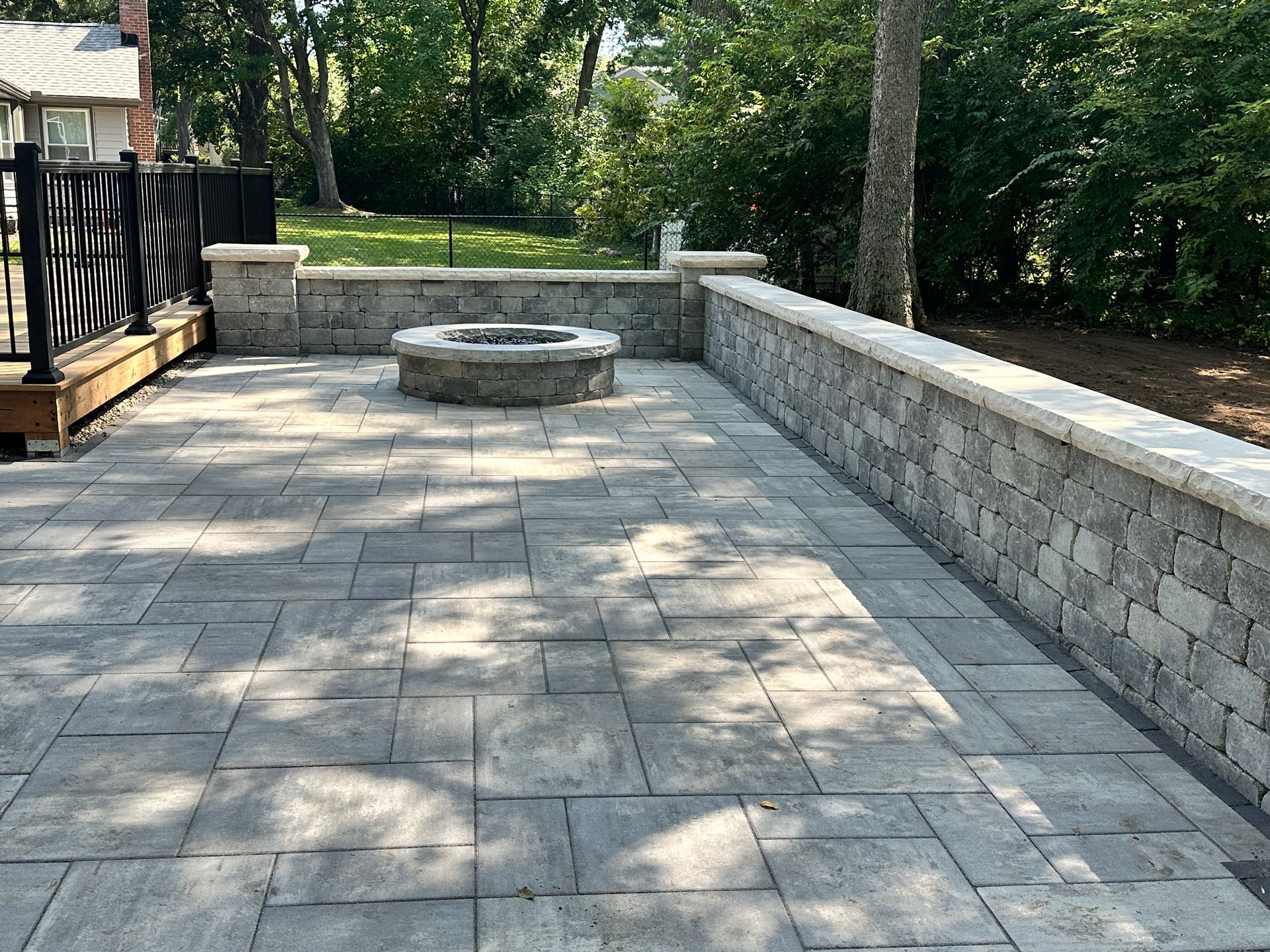 A stone patio featuring a circular fire pit surrounded by a low stone retaining wall in a backyard setting.