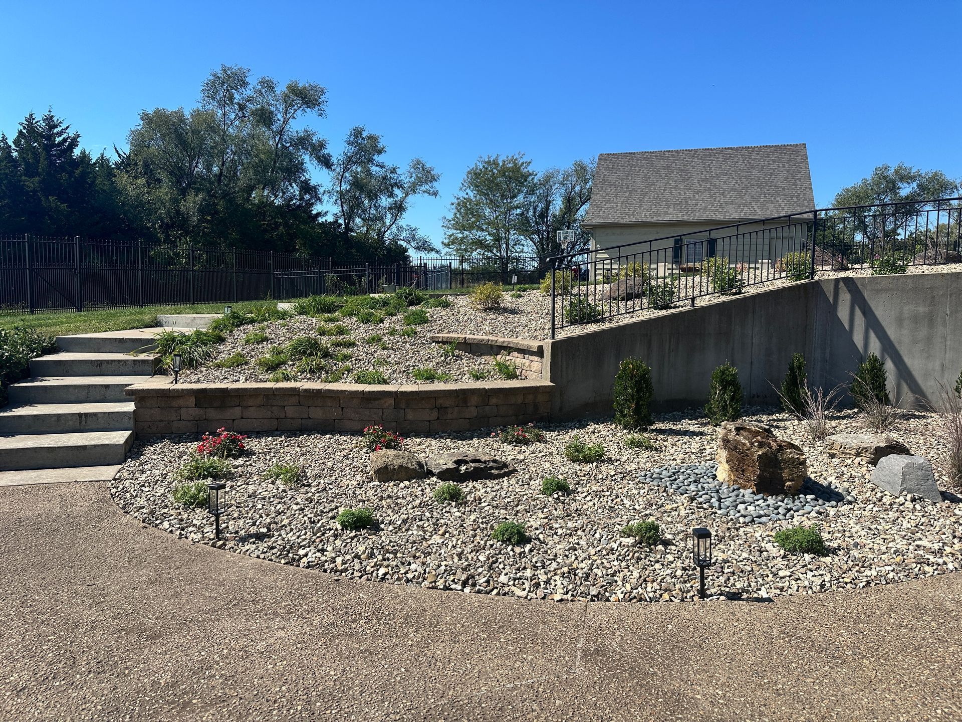 A garden scene with stone stairs leading to a terraced hillside with rocks, small shrubs, and a small building.