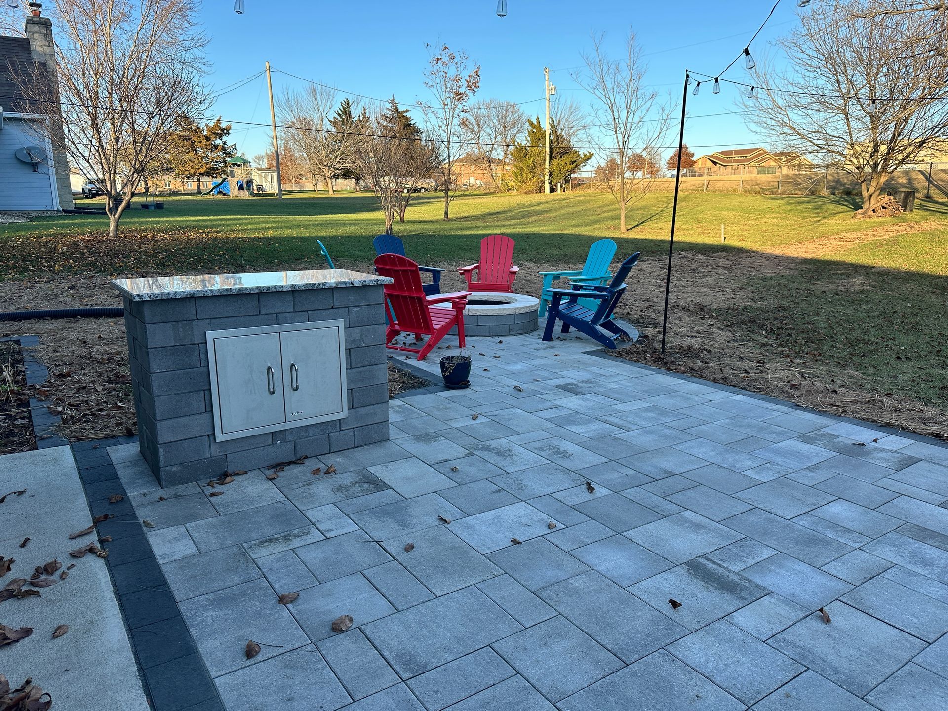 A stone outdoor kitchen island sits on a paved patio with four Adirondack chairs arranged around a fire pit in a yard.