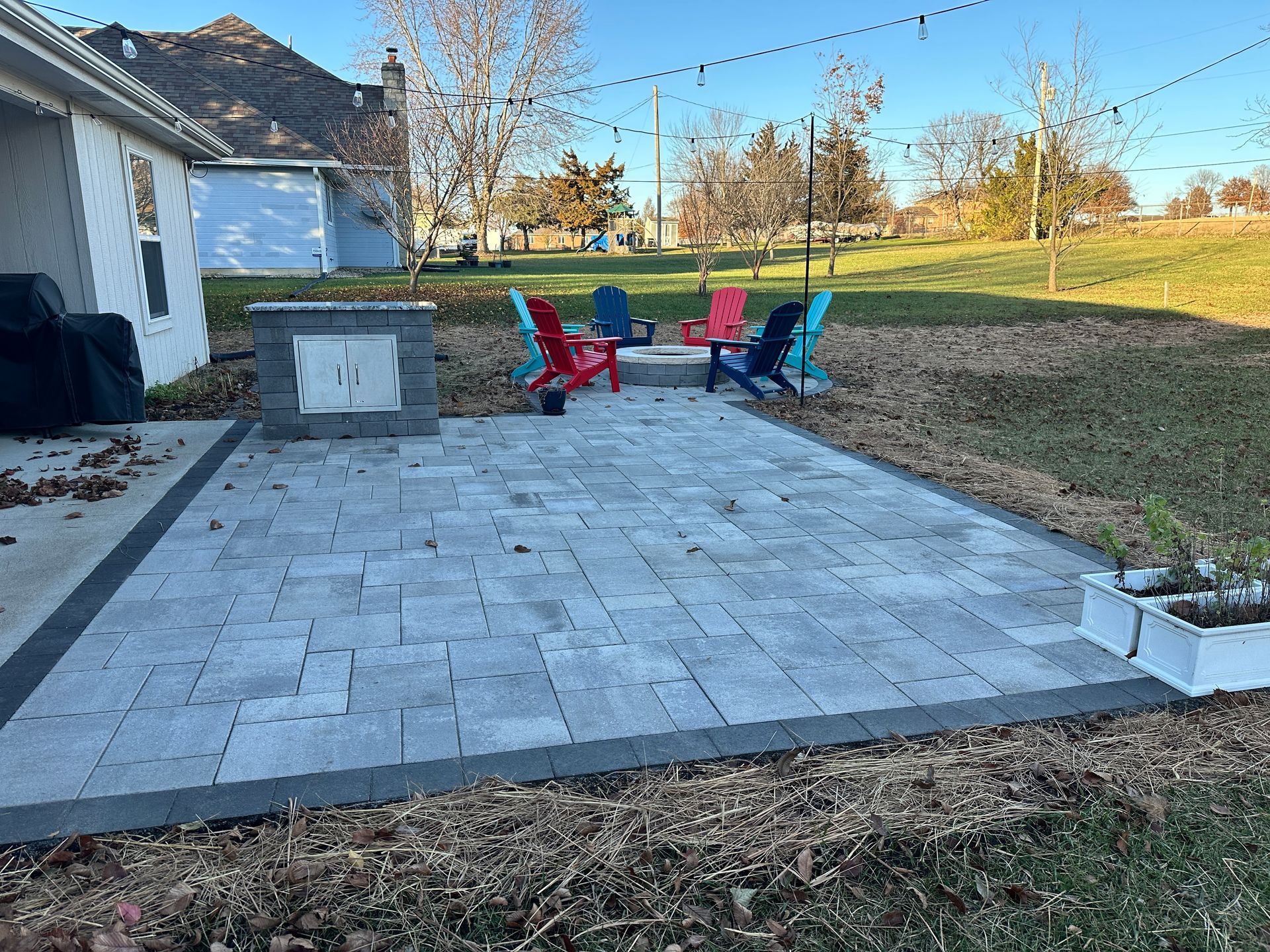 A backyard patio with gray pavers, a stone fire pit, and four colorful Adirondack chairs on a sunny day.