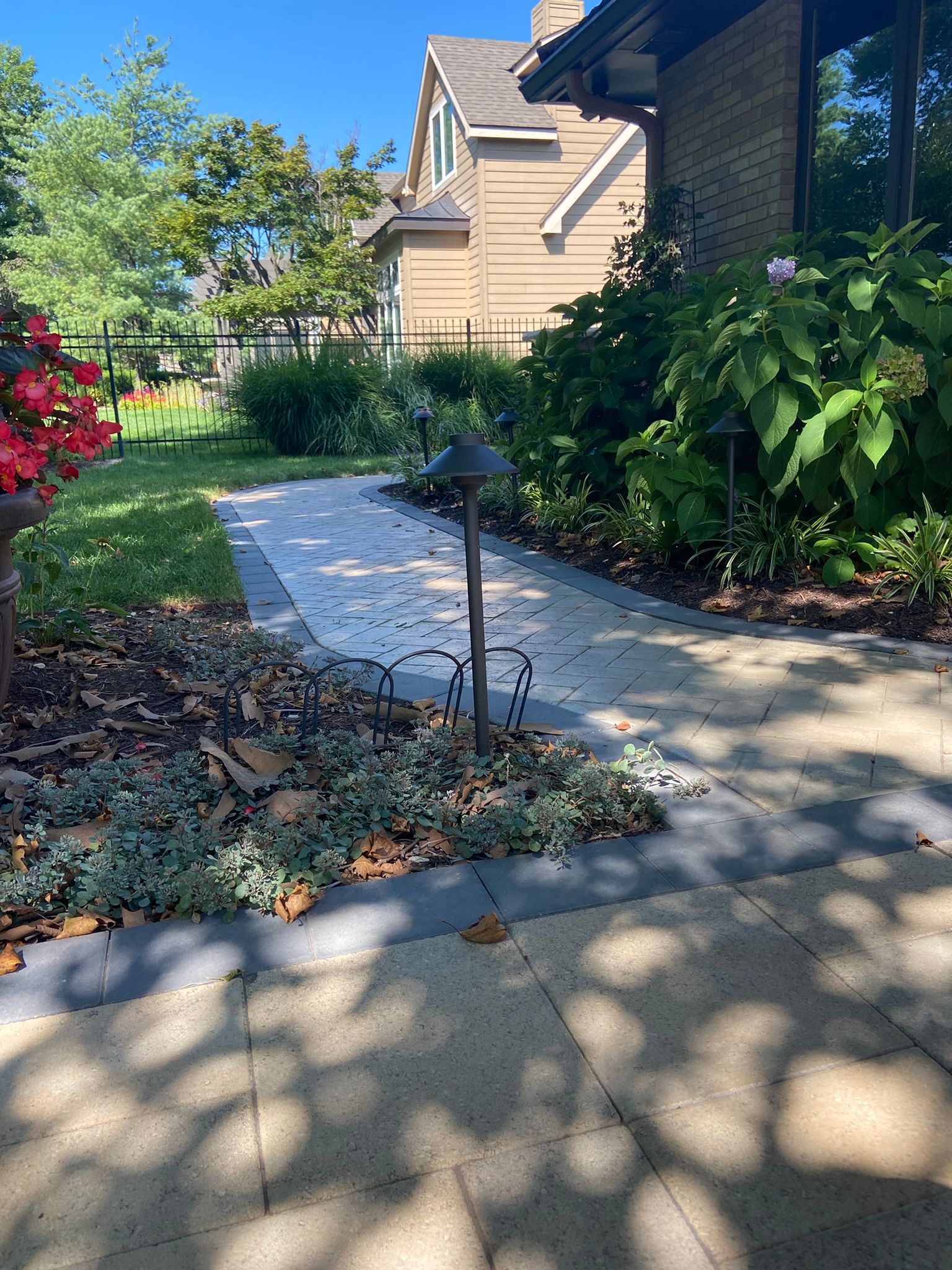 A paved walkway leads to a house, flanked by garden beds with small lights, green plants, and red flowers.