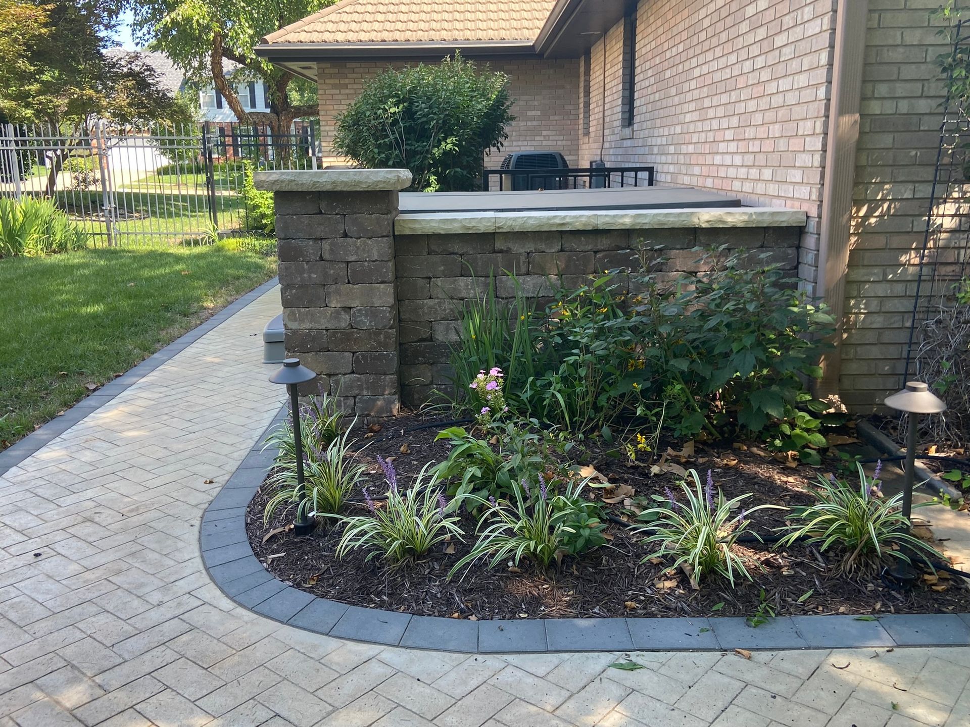 A curved paver walkway leads past a brick planter box with low shrubs toward a house and yard.