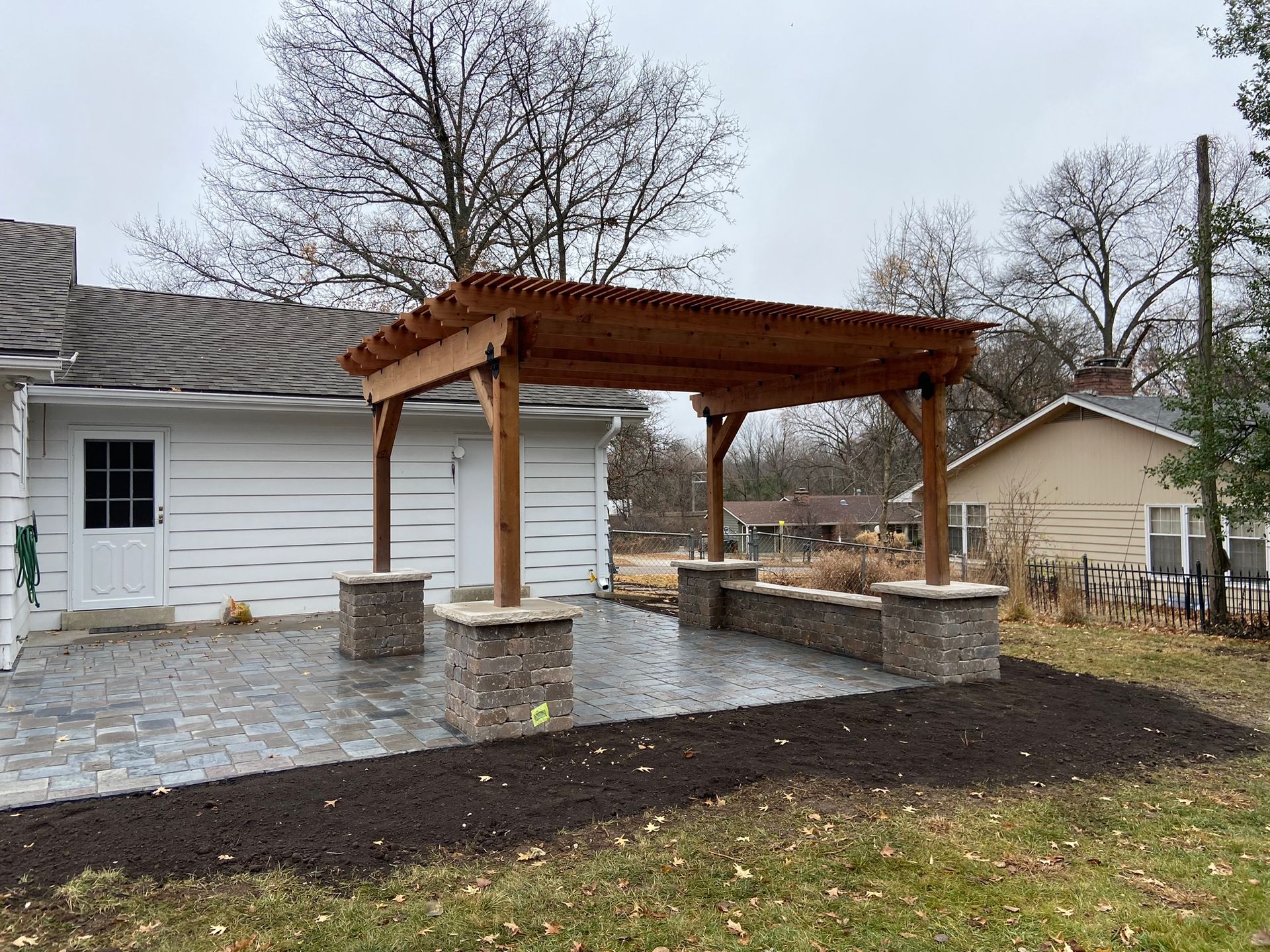 A wooden pergola with stone-base columns sits on a grey paver patio beside a white house in a backyard.