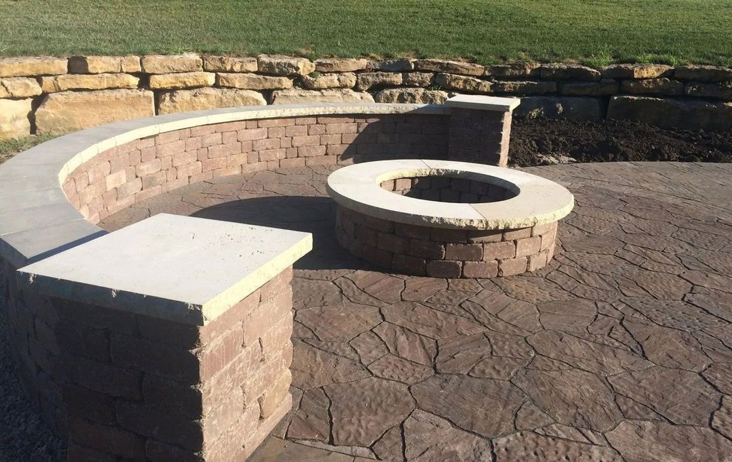 A curved stone seating wall and matching circular fire pit on a patterned patio, with a stone retaining wall in the back.