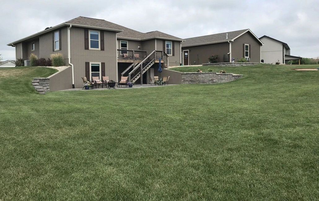 A two-story tan house with a walk-out basement, back deck, and stairs, set on a large grassy lot under a cloudy sky.