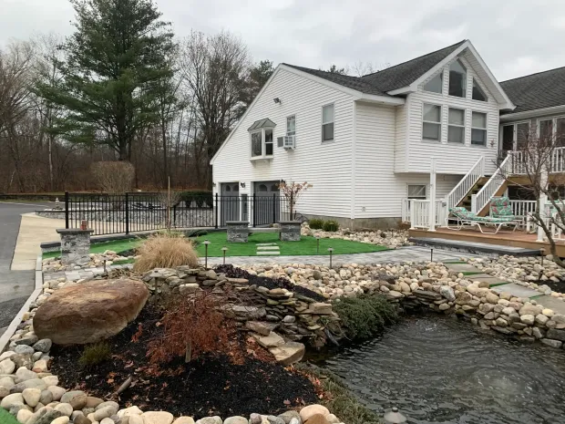 A white multi-story house with a backyard featuring a stone-lined pond, manicured lawn, and a deck.