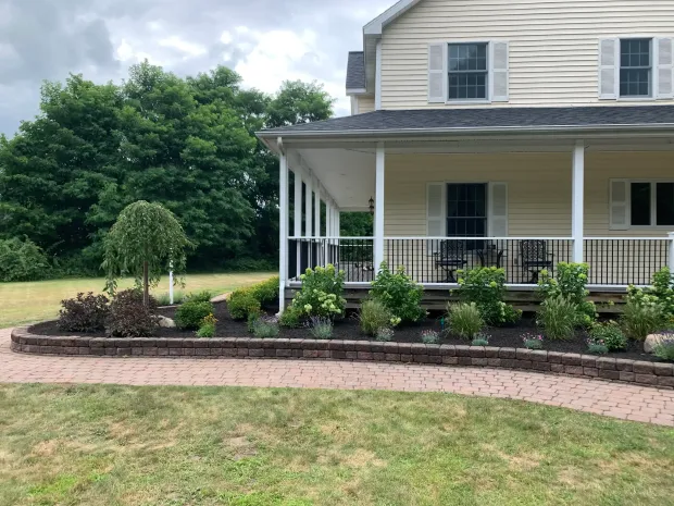 A beige two-story house with a wraparound porch, featuring a stone garden bed in the front and a brick walkway.