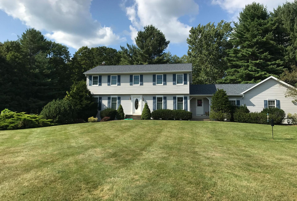 A two-story colonial-style house with light siding and blue shutters, surrounded by green trees and a large lawn.