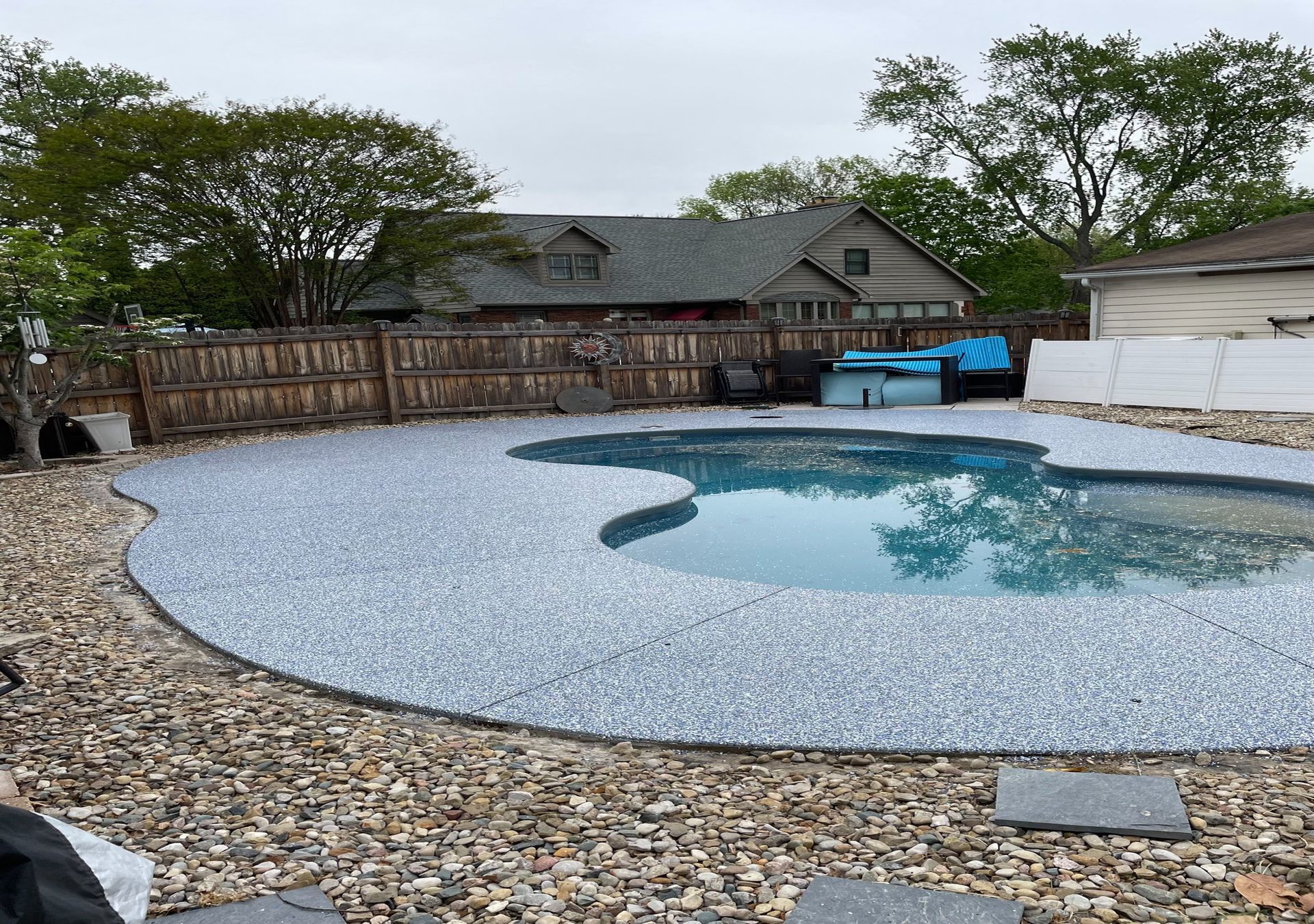 Backyard swimming pool with a pebbled deck, wooden fence, and trees in the background
