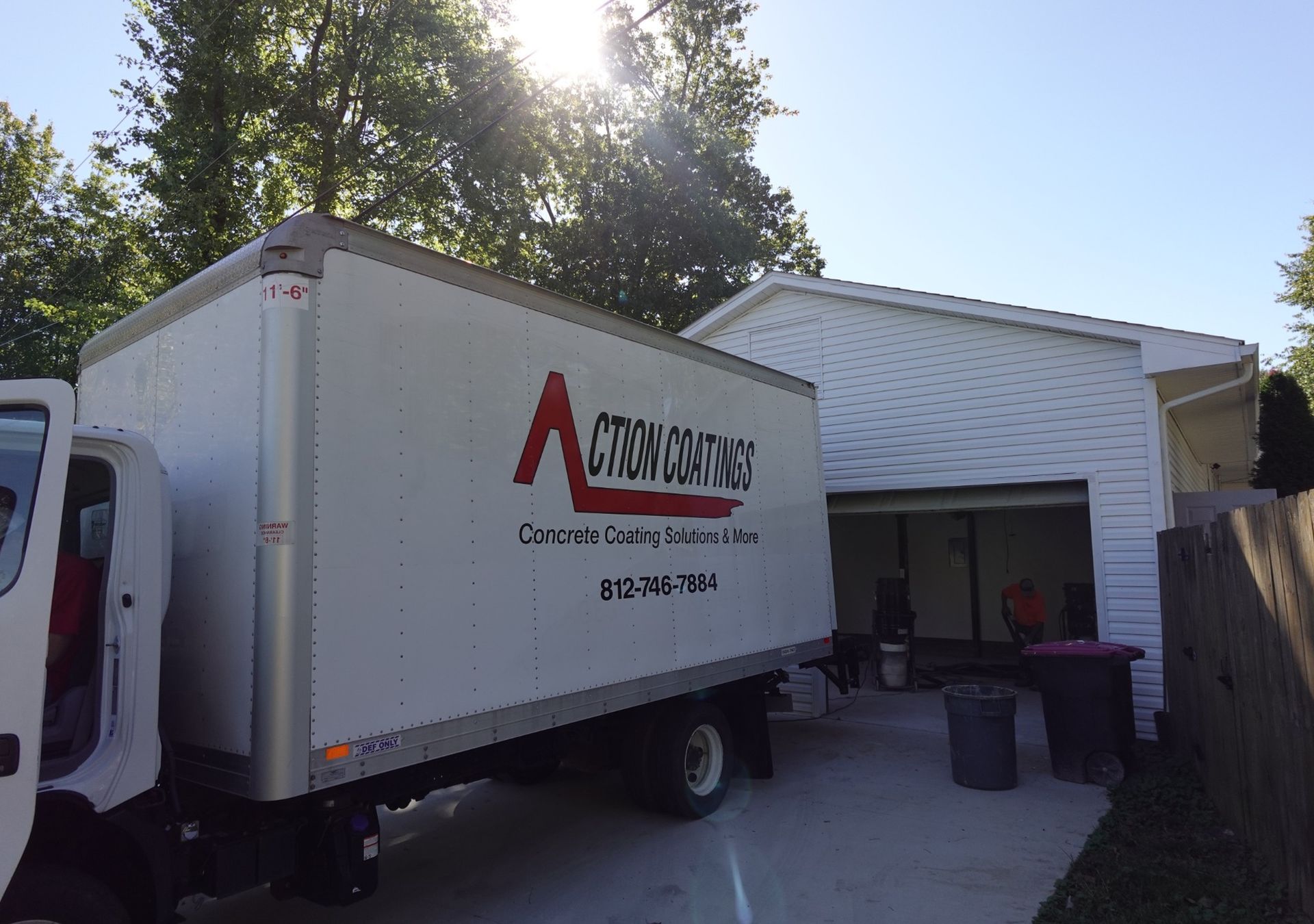 Moving truck parked in a driveway beside a white house, with rear door open and trash bins nearby.