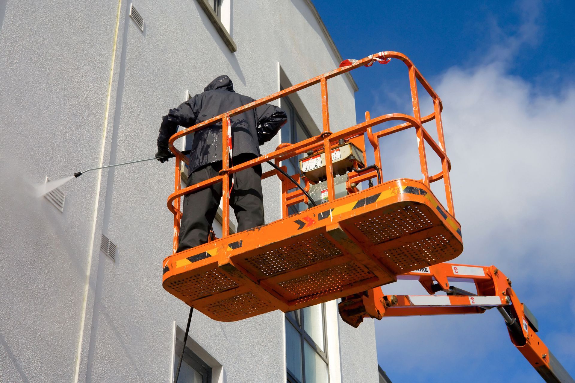 Worker on an orange lift cleaning a building wall with a pressure washer