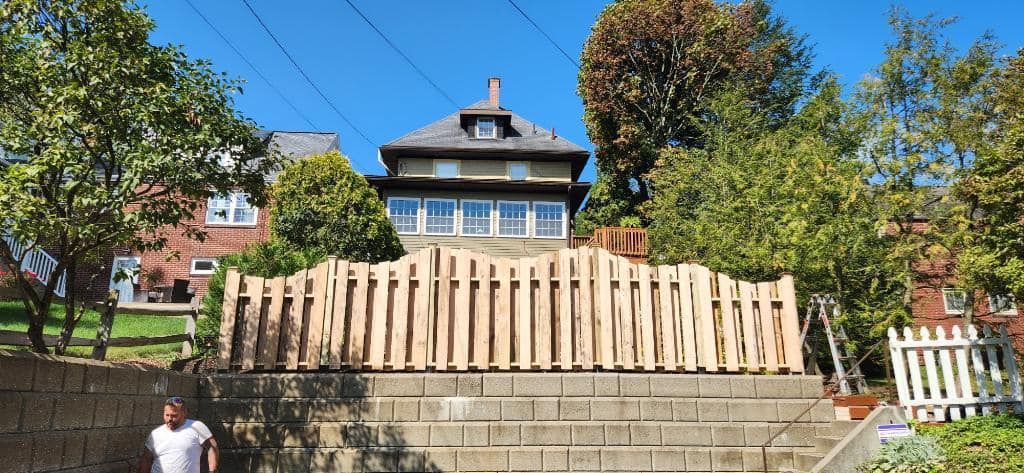 A man is standing in front of a large house with a wooden fence in front of it.