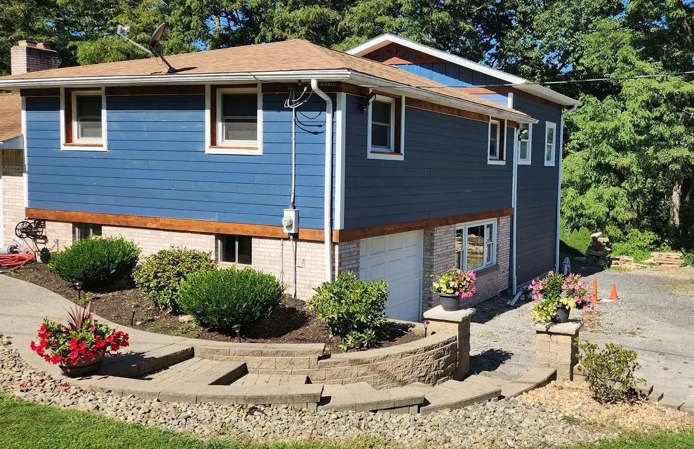 A blue house with a brown roof is surrounded by trees and flowers.