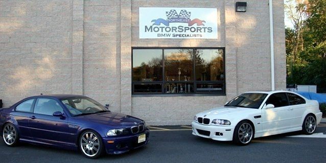 Two BMW M3 coupes parked in front of a Motorsports shop. One is blue, the other white.