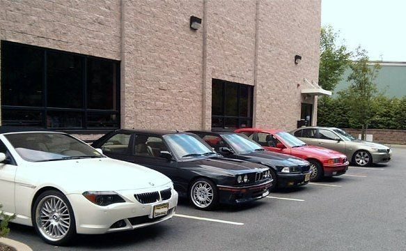 A row of six BMW cars of various models and colors parked in front of a brick building.