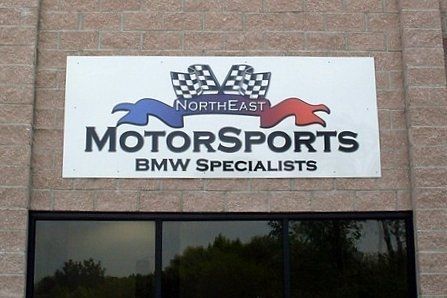 Sign for Northeast Motorsports, BMW specialists. White with logo: checkered flag, blue and red ribbons.