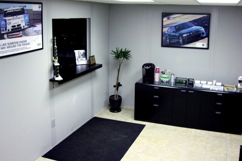 Office interior with service counter, black storage, two car posters, and a small potted plant.