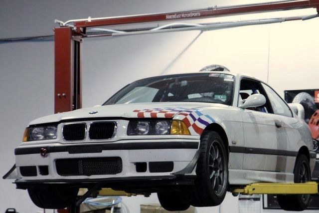 White BMW E36 race car raised on a car lift in a garage, checkered flag design on hood and side.