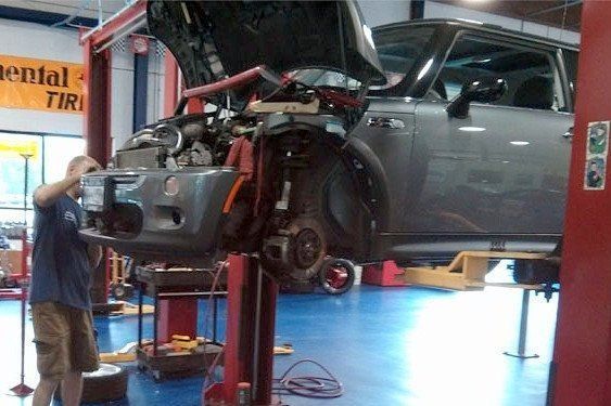 Mechanic working on a gray Mini Cooper raised on a lift in a garage. The hood is up, and the front tire is removed.