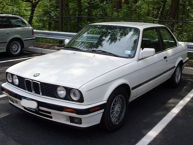 White BMW E30 coupe parked in a lot with a green background.