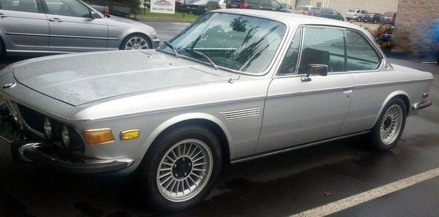 Silver BMW E9 coupe parked on wet pavement.