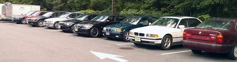 Cars parked in a row on an asphalt surface, trees in the background. Includes black, white, and maroon colored vehicles.