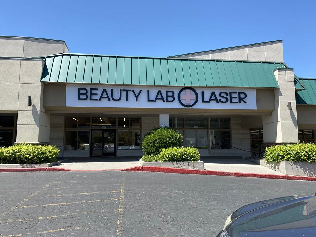 Storefront of "Beauty Lab Laser" with a green metal awning, a sign with a medical cross logo, and green bushes.