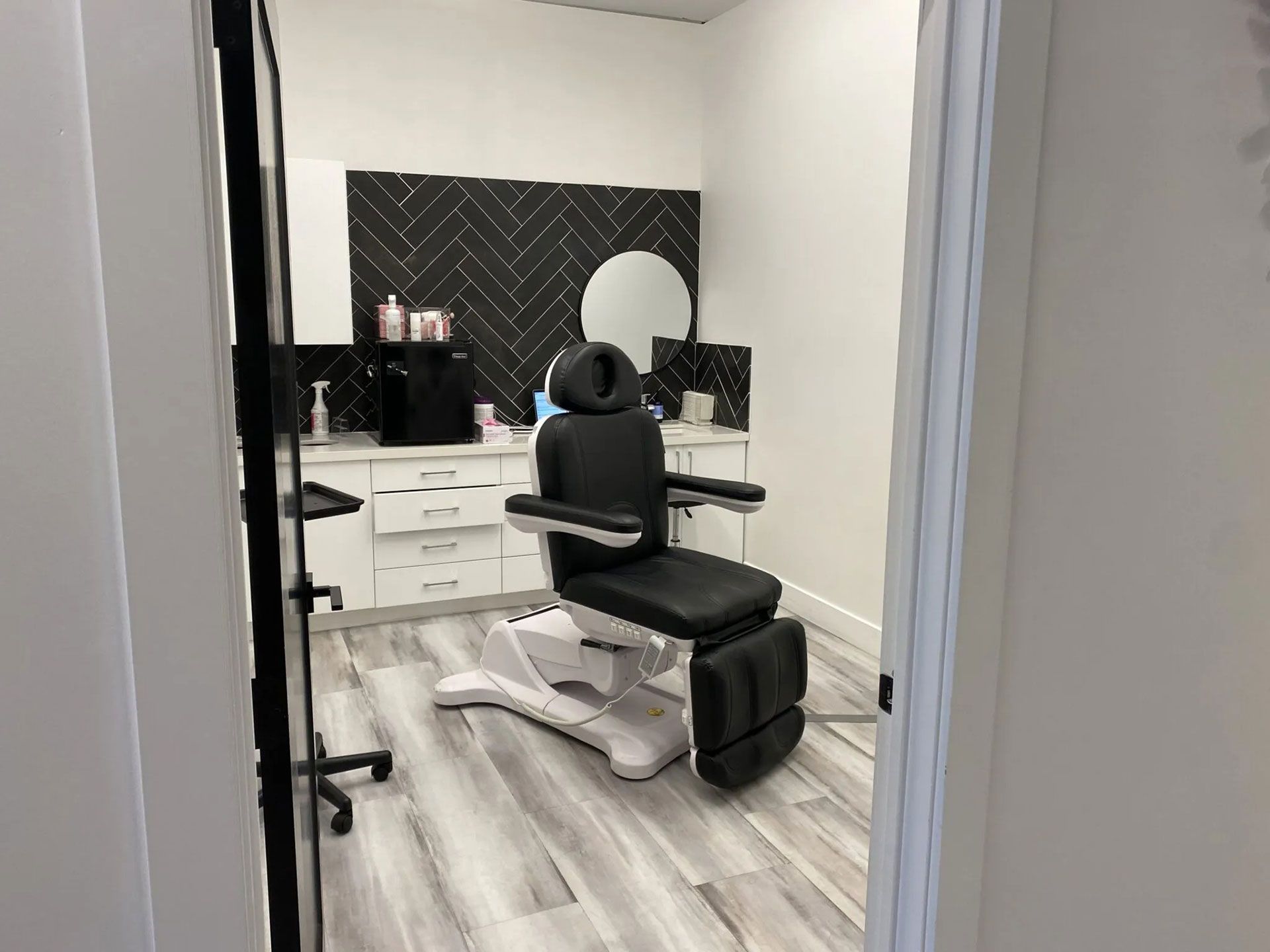 A professional treatment room with a black reclining chair, white cabinets, and a black herringbone tile accent wall.